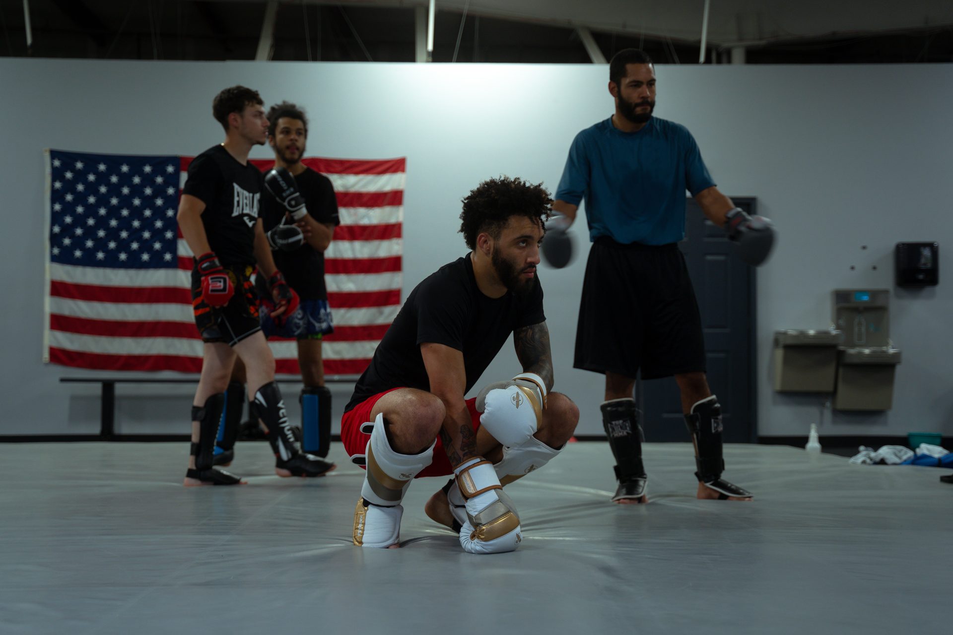 A fighter in gloves and shin pads crouched on the mat between rounds, two more waiting behind him