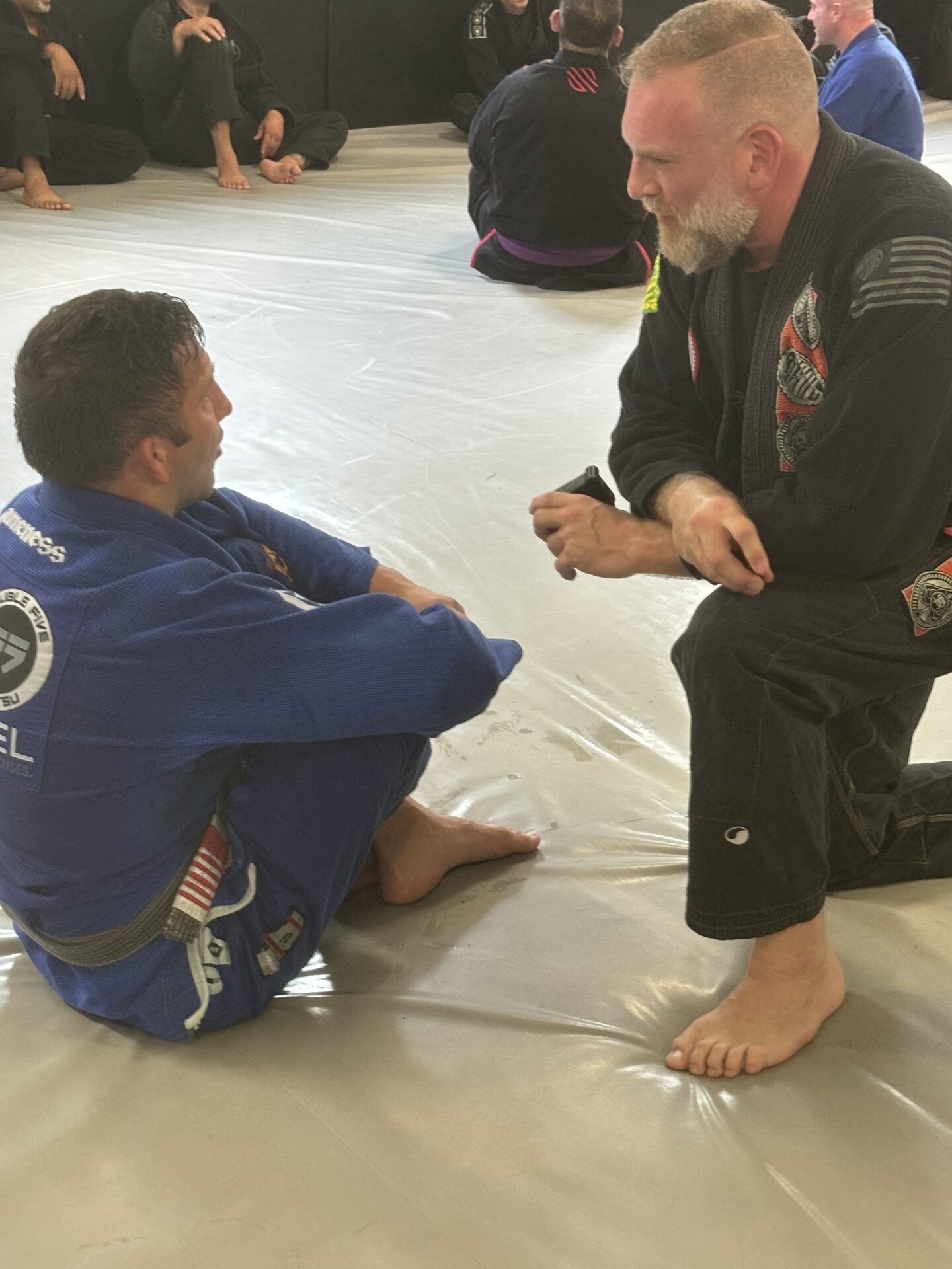 A coach kneeling face to face with one student on the mat, the class seated behind them