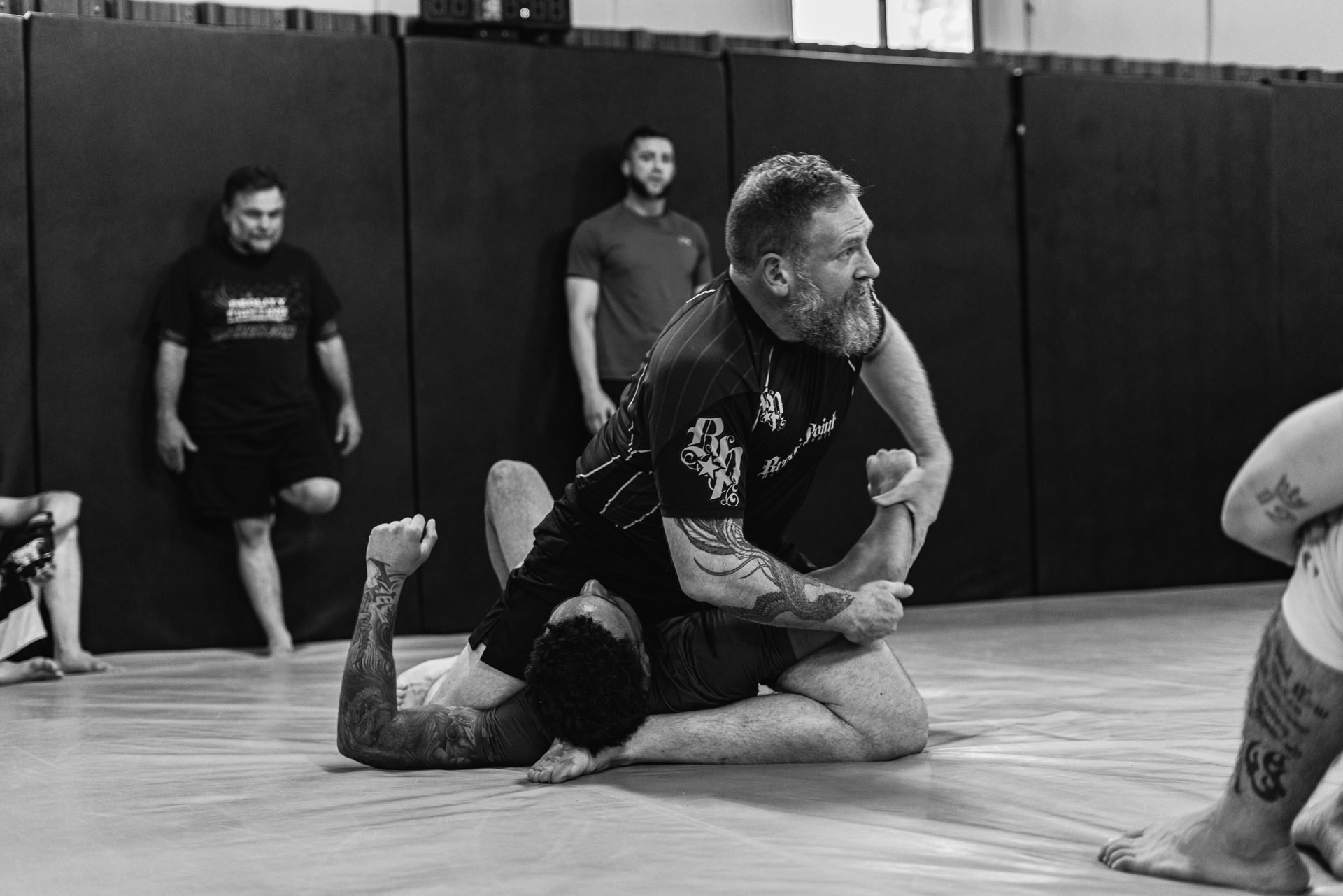 Coach Ruben Gallego holding a top position over a training partner on the mat while the class watches from the wall